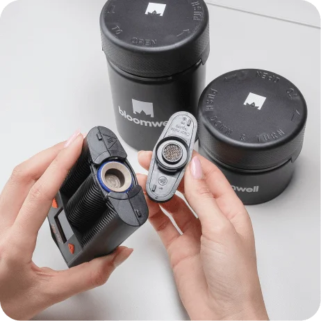 Das Bild zeigt zwei Hände, die einen tragbaren Verdampfer für Cannabis halten. Die linke Hand hält das Hauptgerät, während die rechte Hand ein abnehmbares Teil mit einem Metallsieb hält. Im Hintergrund sind zwei schwarze, zylindrische Behälter mit dem Logo "bloomwell" zu sehen. Die Gerätschaften könnten im Kontext von Cannabis-Rezepten für die medizinische Anwendung genutzt werden.