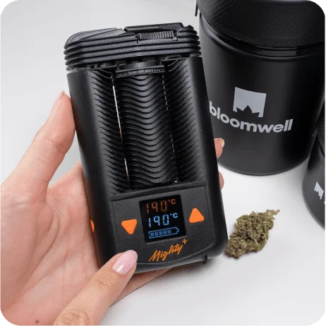 Eine Hand hält einen schwarzen tragbaren Vaporizer, auf dem ein kleines digitales Display mit der Temperaturanzeige "190°C" zu sehen ist. Der Vaporizer hat orangefarbene Bedientasten und den Schriftzug "Mighty+" darauf. Daneben liegt eine kleine Menge getrockneter Cannabisblüten. Im Hintergrund sind schwarze, zylindrische Behälter mit dem Schriftzug "bloomwell" sichtbar. Dieses Bild könnte in einem Kontext stehen, in dem Cannabis für medizinische Rezepte verwendet wird.