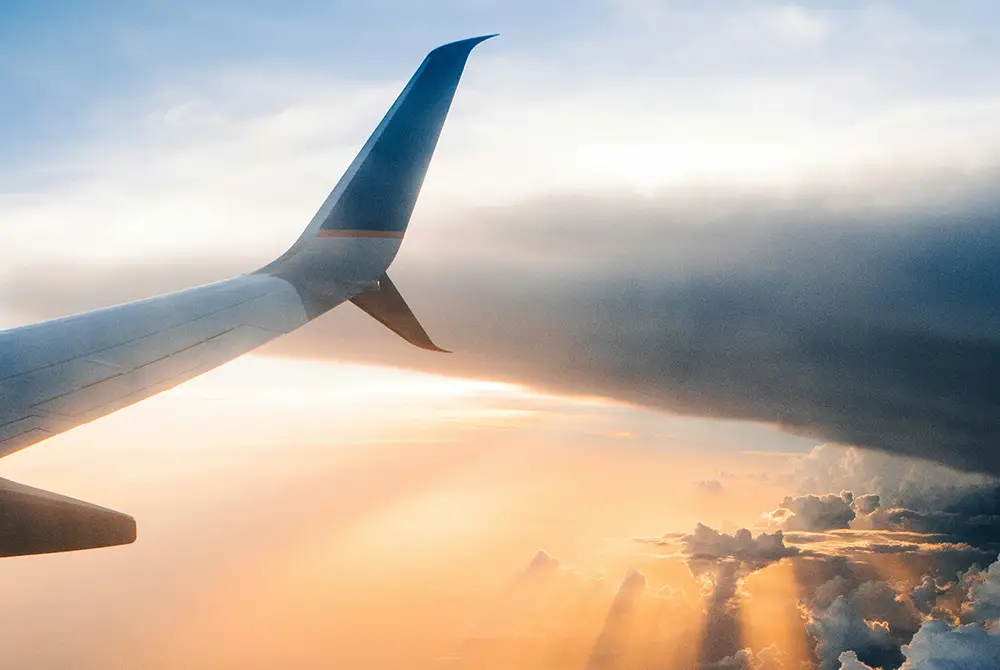 Flugzeugflügel fliegt über Wolken, während Sonnenlicht durch Lücken während des Sonnenuntergangs scheint.