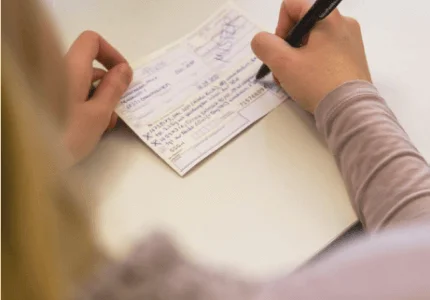 Person, die mit einem schwarzen Stift auf einem Tisch ein Formular oder Dokument ausfüllt.