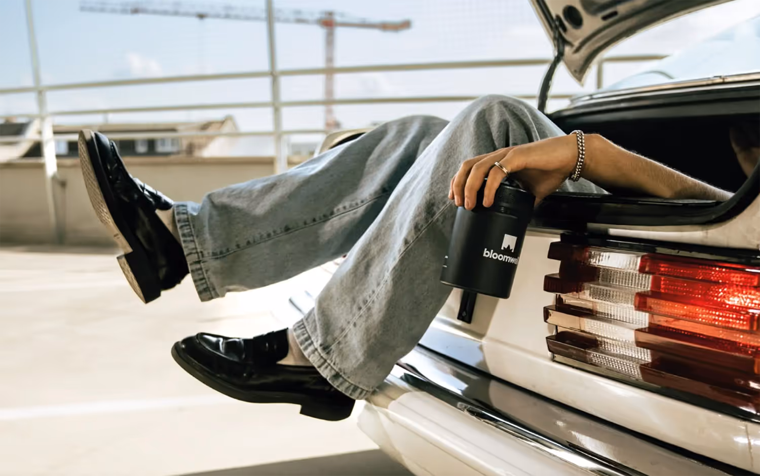 Person, die im offenen Kofferraum eines weißen Autos sitzt, hellblaue Jeans und schwarze Schuhe trägt und eine schwarze Dose mit Bloomwell-Logo in der Hand hält.