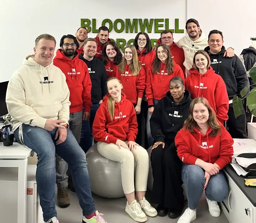 Eine Gruppe von sechzehn Personen in Freizeitkleidung, viele davon mit roten und schwarzen Kapuzenpullis, posiert fröhlich in einem Innenraum vor einem grünen „BLOOMWELL”-Schild an der Wand.