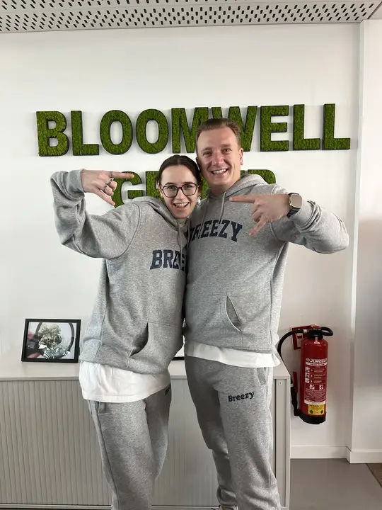 Zwei lächelnde Personen in passenden grauen Breezy-Hoodies und Jogginghosen posieren mit Handzeichen vor einer Wand mit grünen Moosbuchstaben, die das Wort BLOOMWELL bilden.