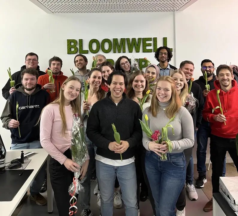 Eine Gruppe lächelnder junger Erwachsener mit gelben Tulpenblumen in den Händen steht vor einer Wand mit der Aufschrift „BLOOMWELL CARE”.