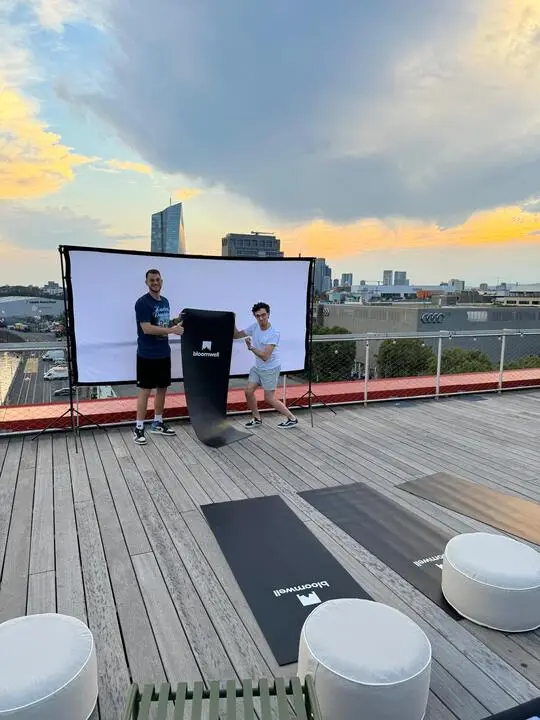 Zwei Personen halten eine schwarze Bloomwell-Yogamatte auf einem Dach mit Stadtgebäuden im Hintergrund während des Sonnenuntergangs.