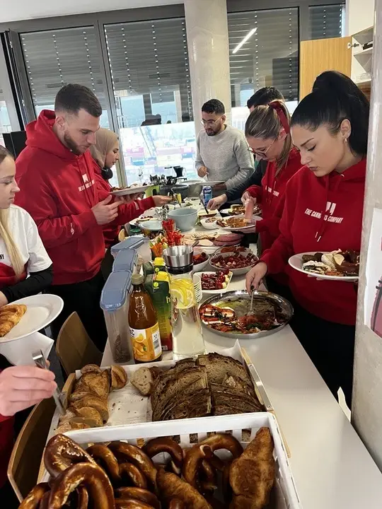 Eine Gruppe von Menschen bedient sich an einem Buffettisch mit Brot, Brezeln, Croissants und verschiedenen Speisen in einem hellen Raum.