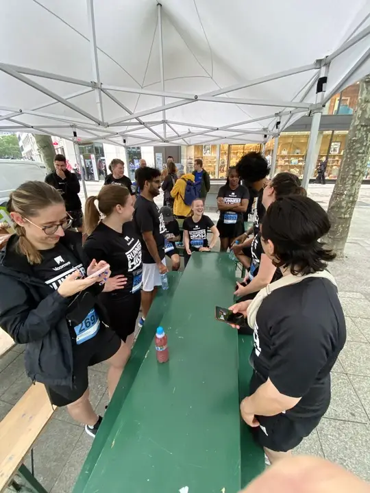 Eine Gruppe von Läufern in schwarzen Event-Shirts versammelte sich unter einem weißen Zelt um einen grünen Tisch, unterhielt sich und schaute auf ihre Handys.