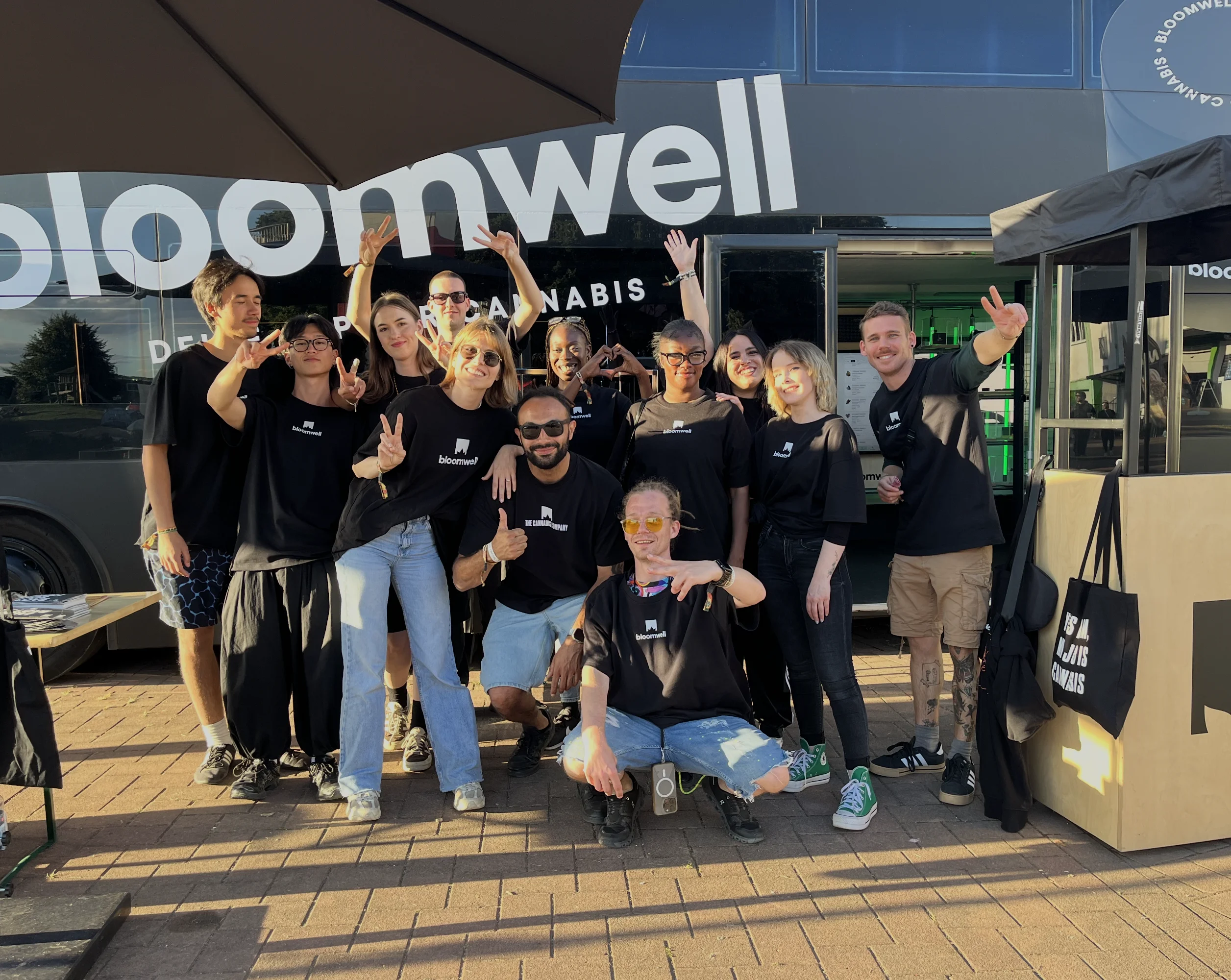 Eine Gruppe junger Leute in schwarzen Bloomwell-Shirts posiert fröhlich vor einem Bloomwell-Cannabis-Truck.
