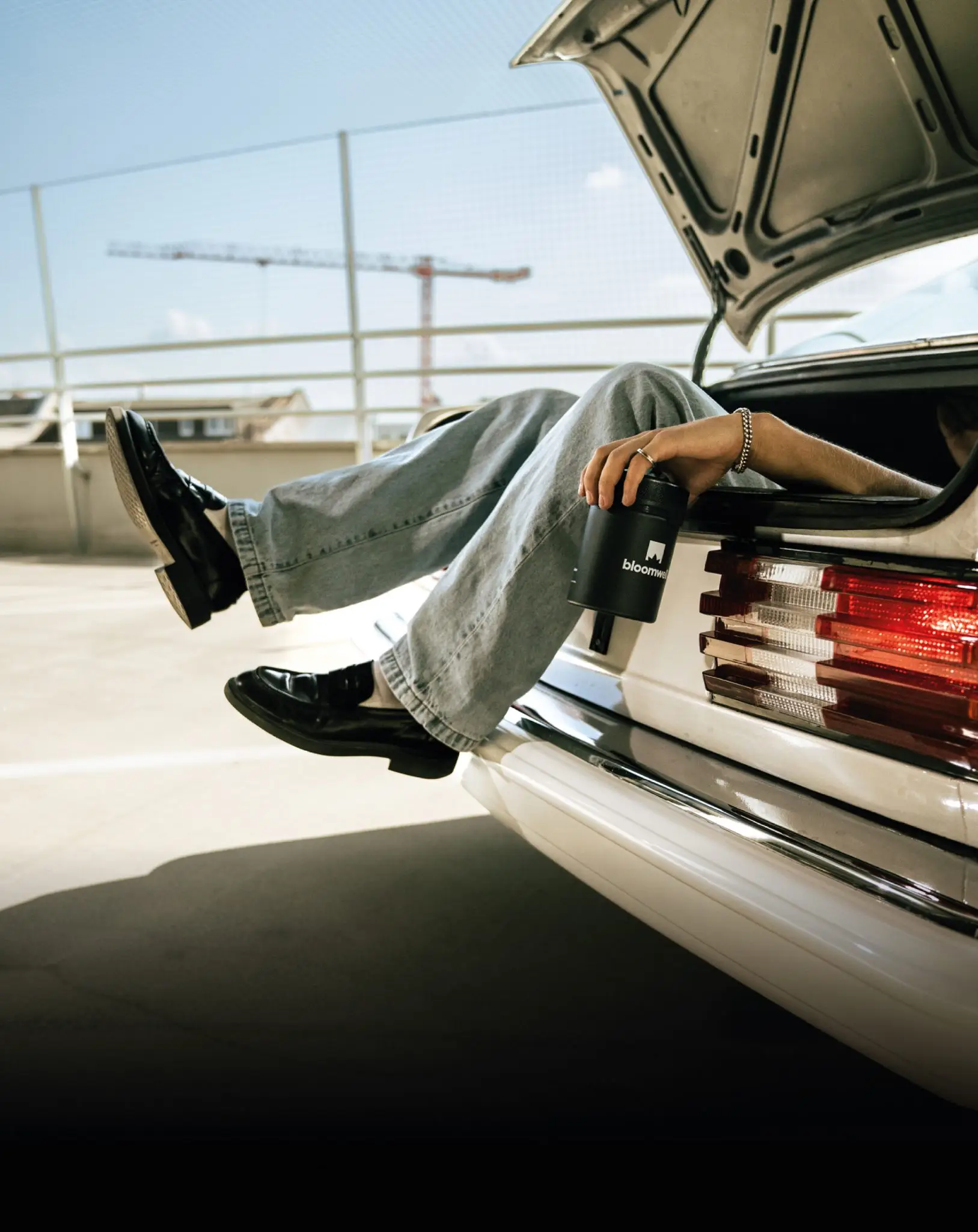 Beine in einer Jeans baumeln aus dem Kofferraum von einem alten vintage Mercedes. In der Hand hält der Mensch eine Blommwell Dose.