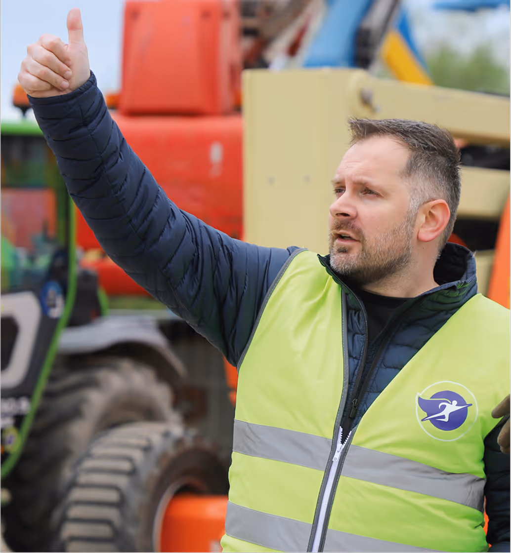 Homme en gilet jaune avec pouce levé devant des machines de chantier.