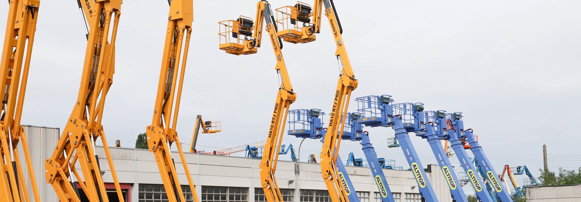 Des plateformes élévatrices jaunes et bleues alignées devant un bâtiment industriel.