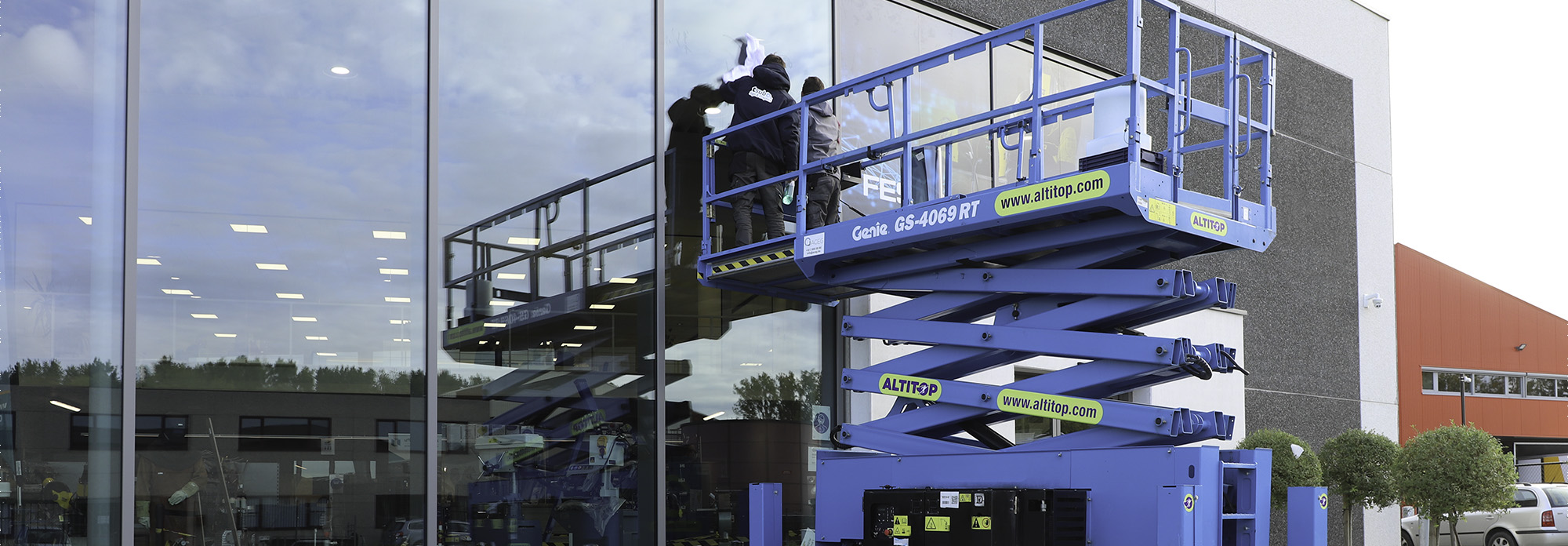 Deux ouvriers nettoient une grande vitre en hauteur avec une plateforme élévatrice bleue.
