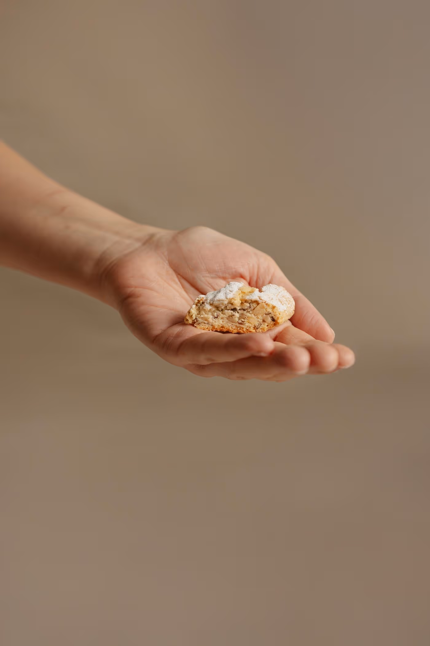 A light-skinned hand holds a small, powdered sugar-dusted cookie with visible nuts, against a soft beige background.