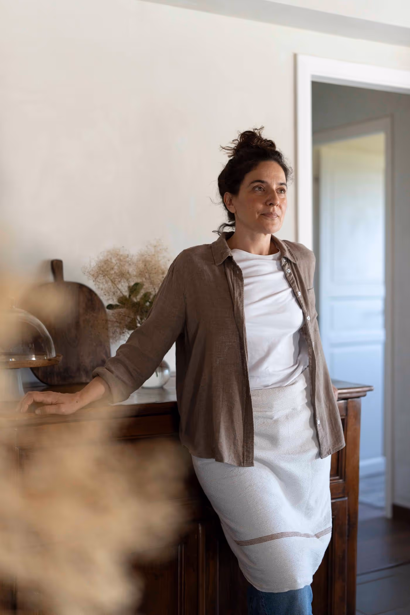 Barbara Grimoldi in a brown linen shirt and white apron leans against a dark wooden cabinet with her hand, looking thoughtfully to the side. Dried flowers and a cutting board are visible on the cabinet.