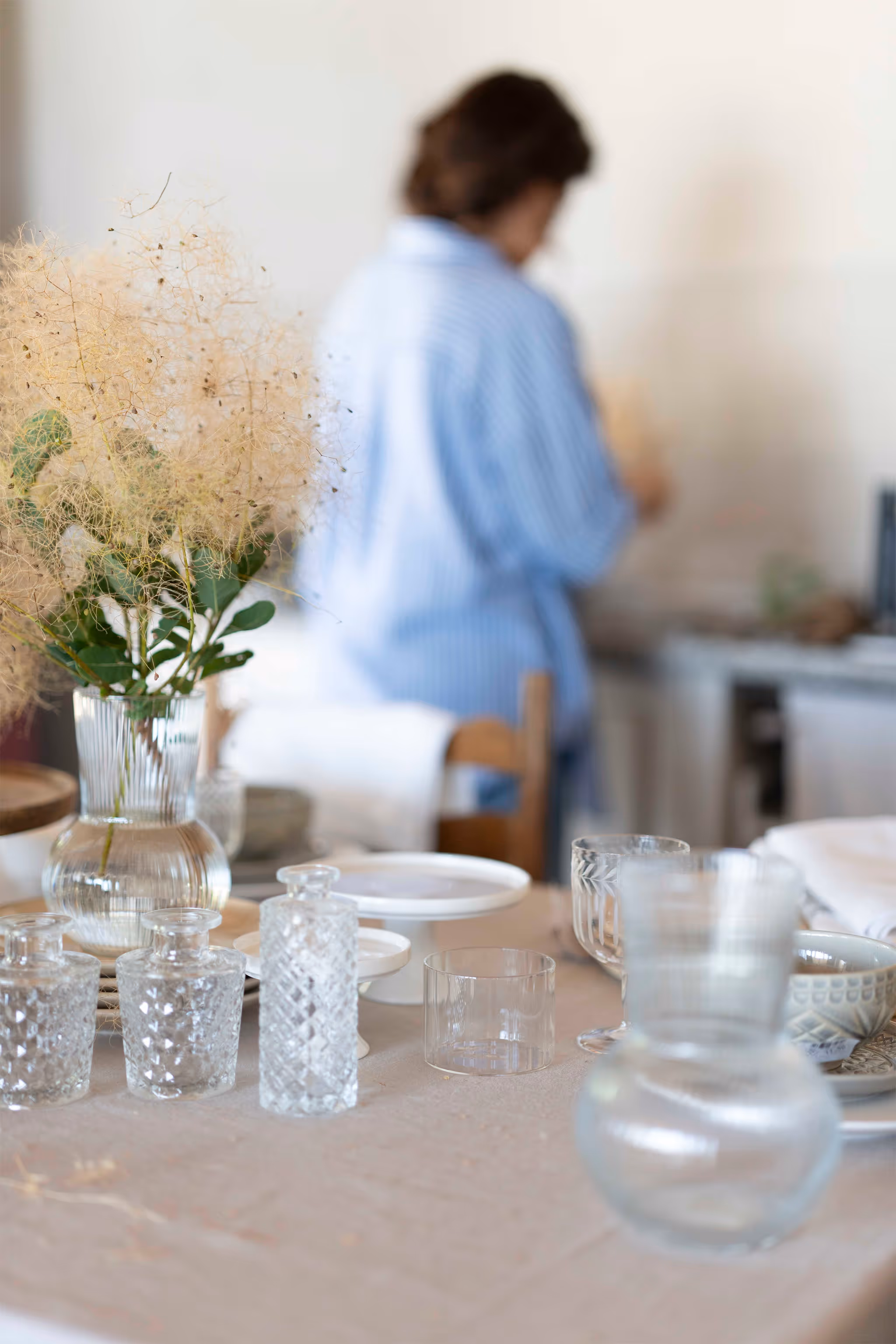 A rustic table set with vintage-style clear glass drinkware and a vase of feathery smoke bush, with a person in a blue and white striped shirt out of focus in the background.