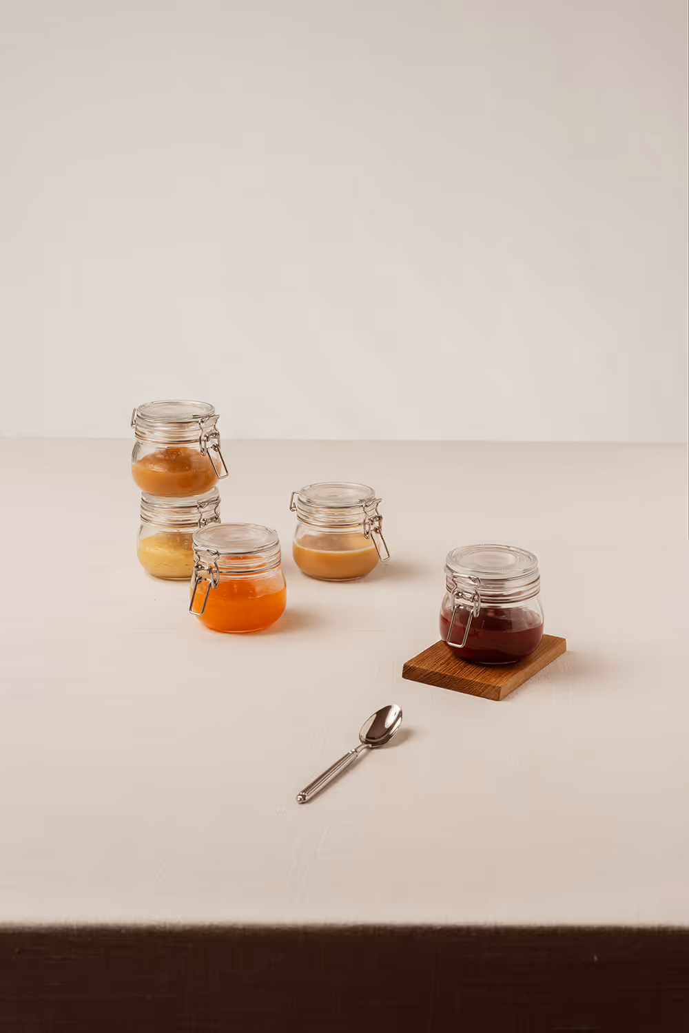 Five small glass jars containing various colored jams arranged on a beige tablecloth, with a silver spoon placed in front of them.