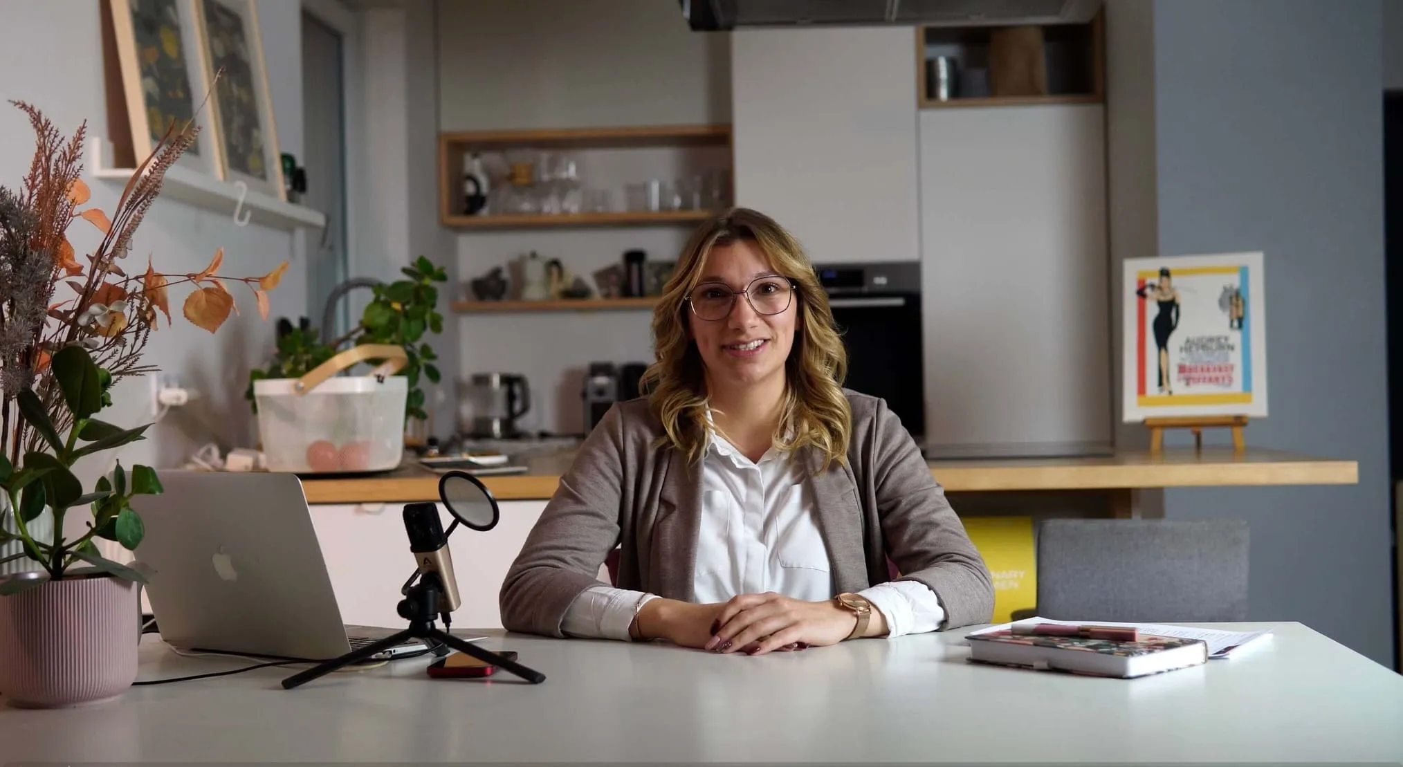 Kobieta (Sandra Kos) z blond włosami i okularami siedzi za stołem z laptopem, mikrofonem i książkami w nowoczesnej kuchni.