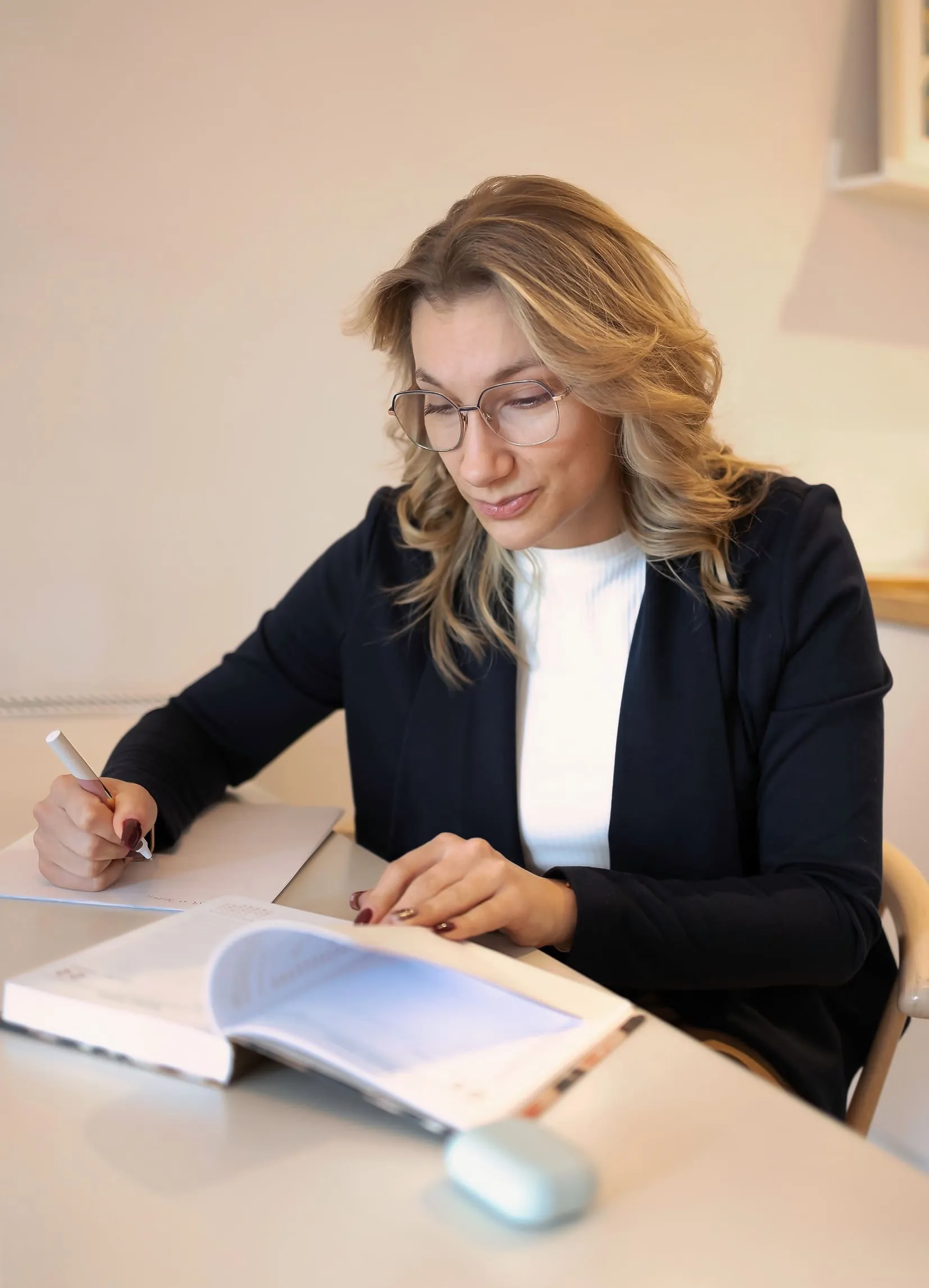 Kobieta (Sandra Kos) w okularach pisze w kalendarzu, przeglądając go na stole.