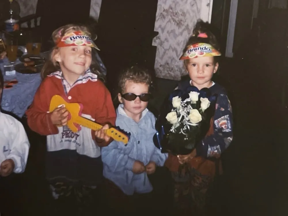 Trzy dziewczynki pozują na zdjęciu, jedna dziewczynka trzyma żółtą zabawkową gitarę, druga dziewczynka (Sandra Kos) w okularach przeciwsłonecznych stoi obok, a trzecia dziewczynka z czapką z daszkiem trzyma bukiet białych róż.