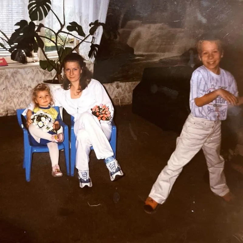 Trzy osoby w pokoju: kobieta i mała dziewczynka siedzą na niebieskich krzesłach trzymając po bukietach kwiatów, chłopiec sto obok i i uśmiecha się.