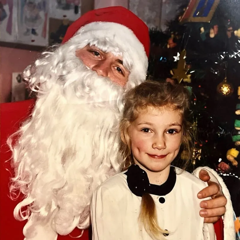 Mężczyzna przebrany za Świętego Mikołaja z białą brodą i czerwoną czapką z ręką na ramieniu uśmiechającej się dziewczynki z blond warkoczem i białą koszulą na tle udekorowanej choinki.