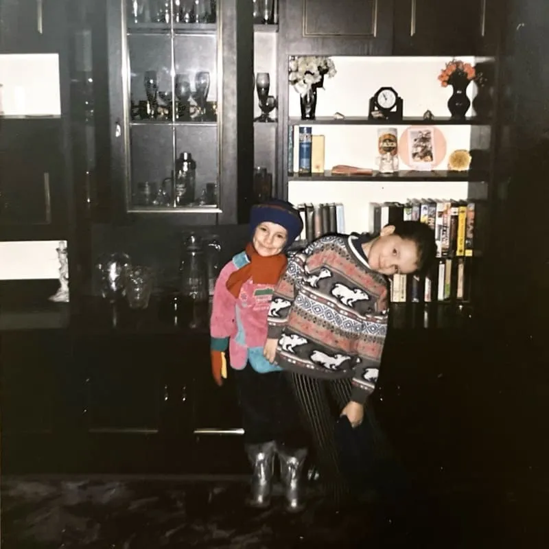 Dwoje dzieci ubrane w kolorowe ubrania i zabawne swetry stoi razem w ciemnym pokoju z witrynką i półką pełną książek i dekoracji.