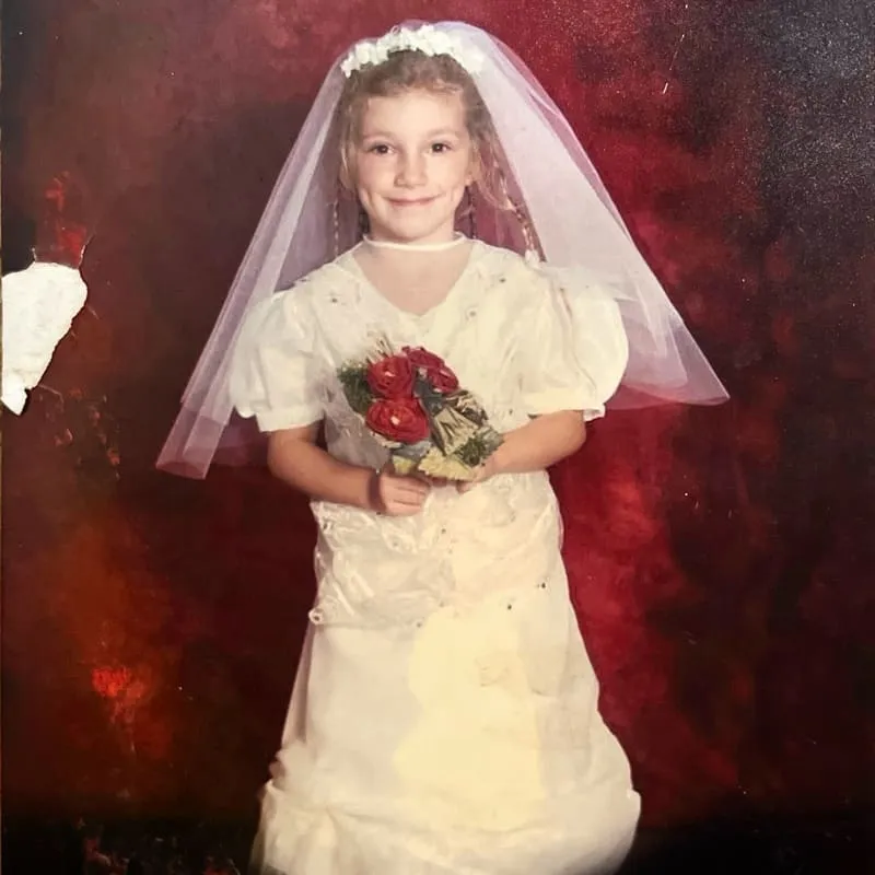Mała dziewczynka uśmiechająca się, ubrana w białą suknię komunijną i welon, trzymająca bukiet czerwonych kwiatów.