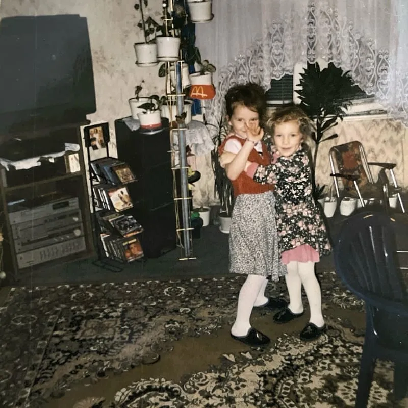 Dwie dziewczynki w sukienkach tańczą razem w salonie z kwiecistym dywanem i roślinami doniczkowymi.