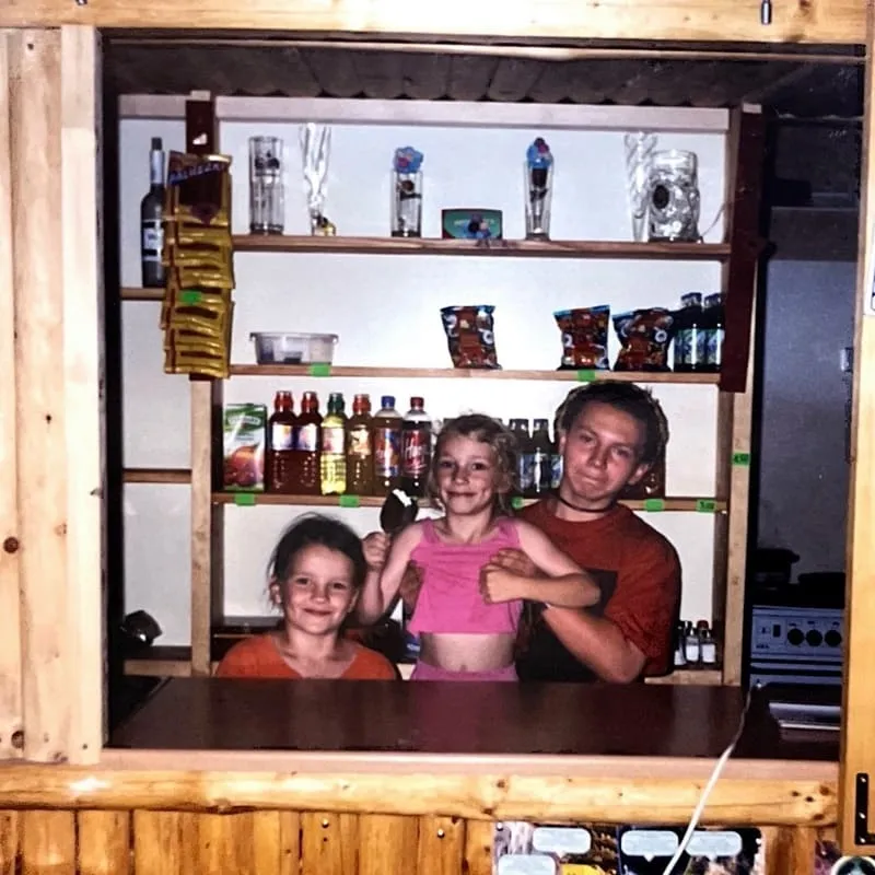 Trójka dzieci za ladą sklepową z półkami pełnymi napojów i przekąsek w tle.