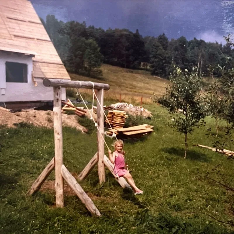 Dziewczynka w różowym ubraniu siedzi na huśtawce zbudowanej z drewnianych kłód w trawiastym ogrodzie obok drewnianego domu i drzew.