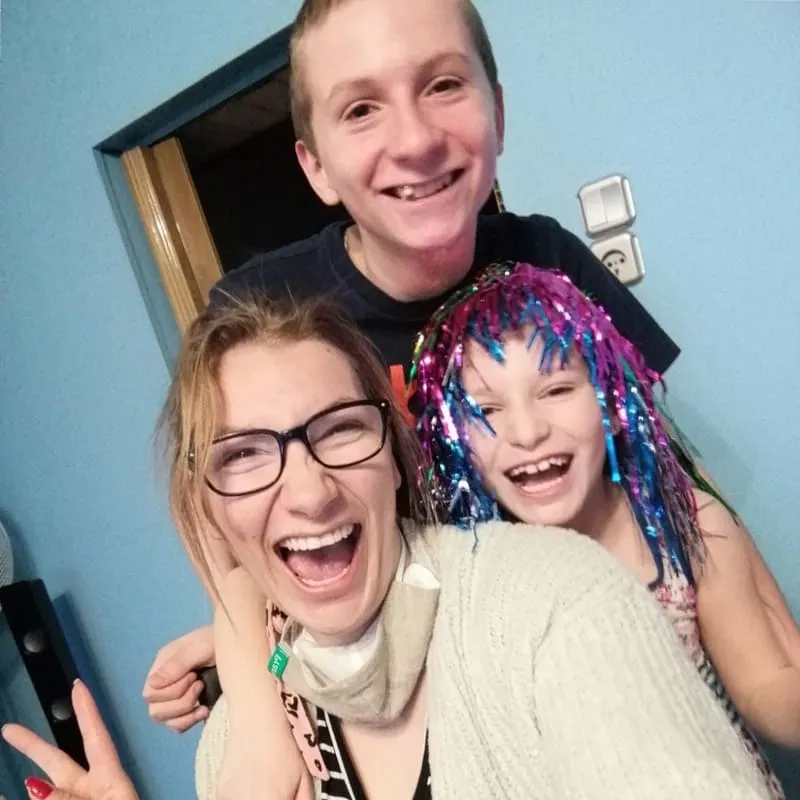 Trzy osoby śmiejące się, w tym kobieta w okularach (Sandra Kos), chłopiec i dziewczynka w kolorowej peruce, pozujące do zdjęcia wewnątrz pomieszczenia.