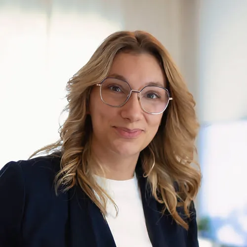 Kobieta w okularach i jasnobrązowych, falowanych włosach, ubrana w biały top i ciemną marynarkę, uśmiechająca się na rozmytym jasnym tle.