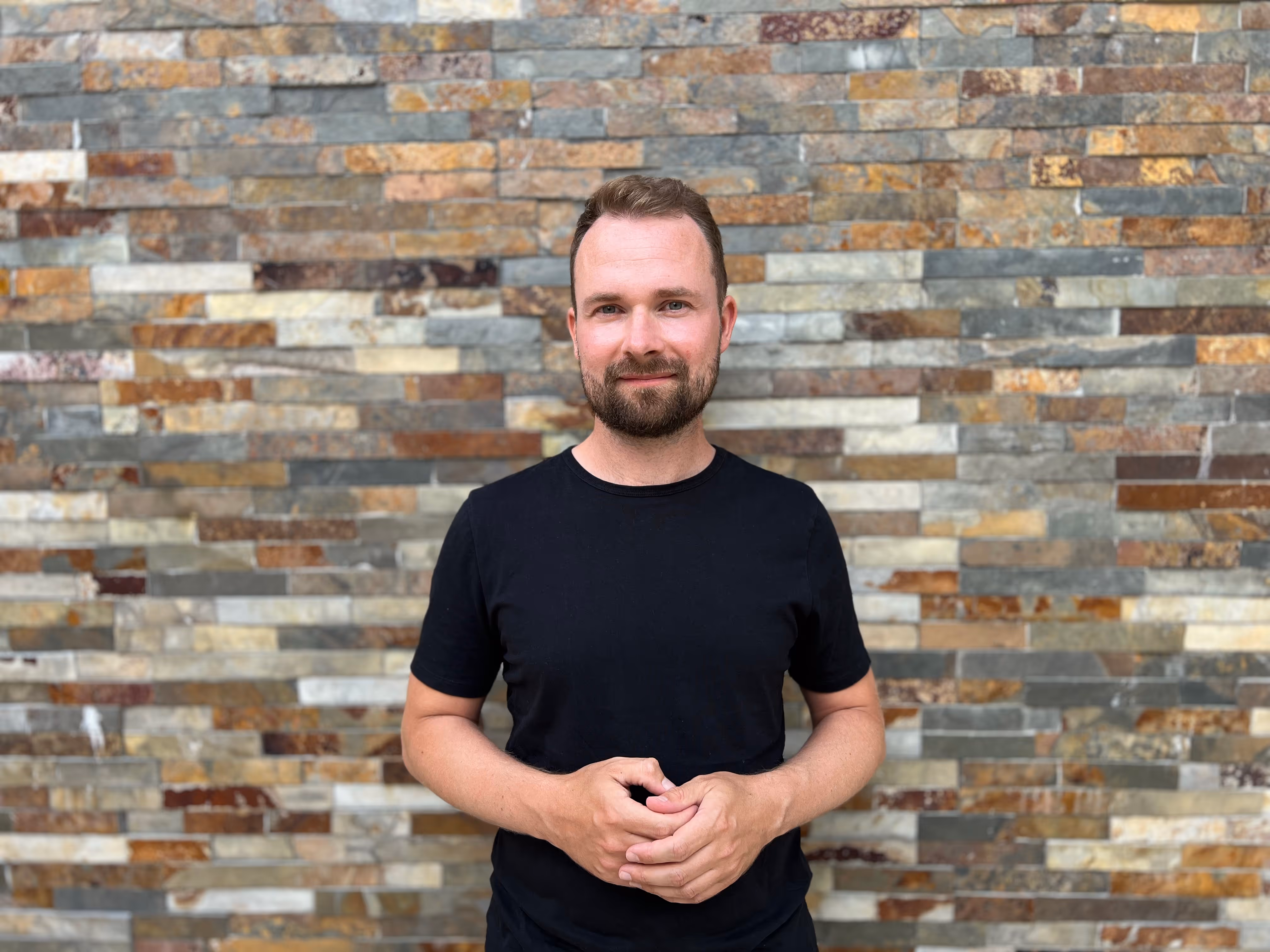 Mann mit kurzem braunem Haar und Bart, der ein schwarzes T-Shirt trägt und vor einer mehrfarbigen Steinmauer steht.