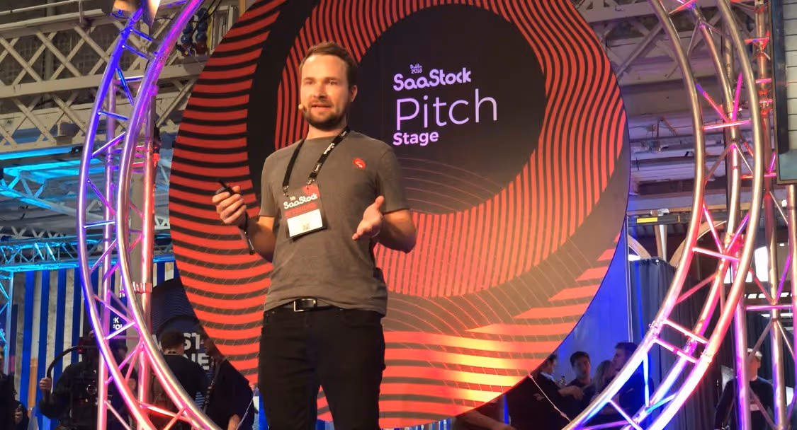 Mann hält eine Präsentation auf der SaaStock Pitch Stage mit einem großen roten und schwarzen runden Hintergrund.