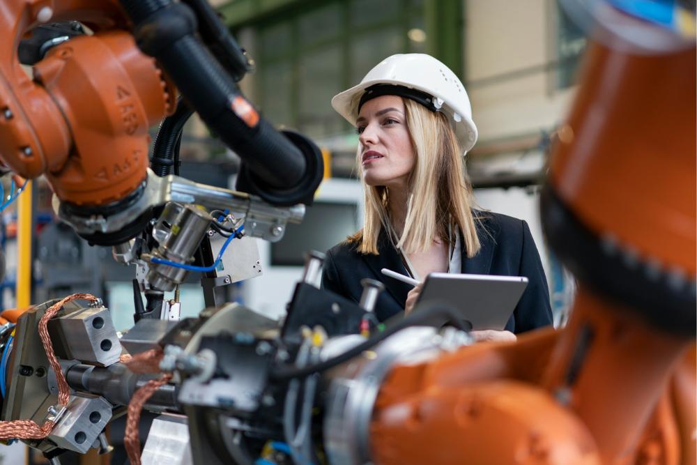 Frau mit Schutzbrille beobachtet Roboterarme in einer Fertigungshalle