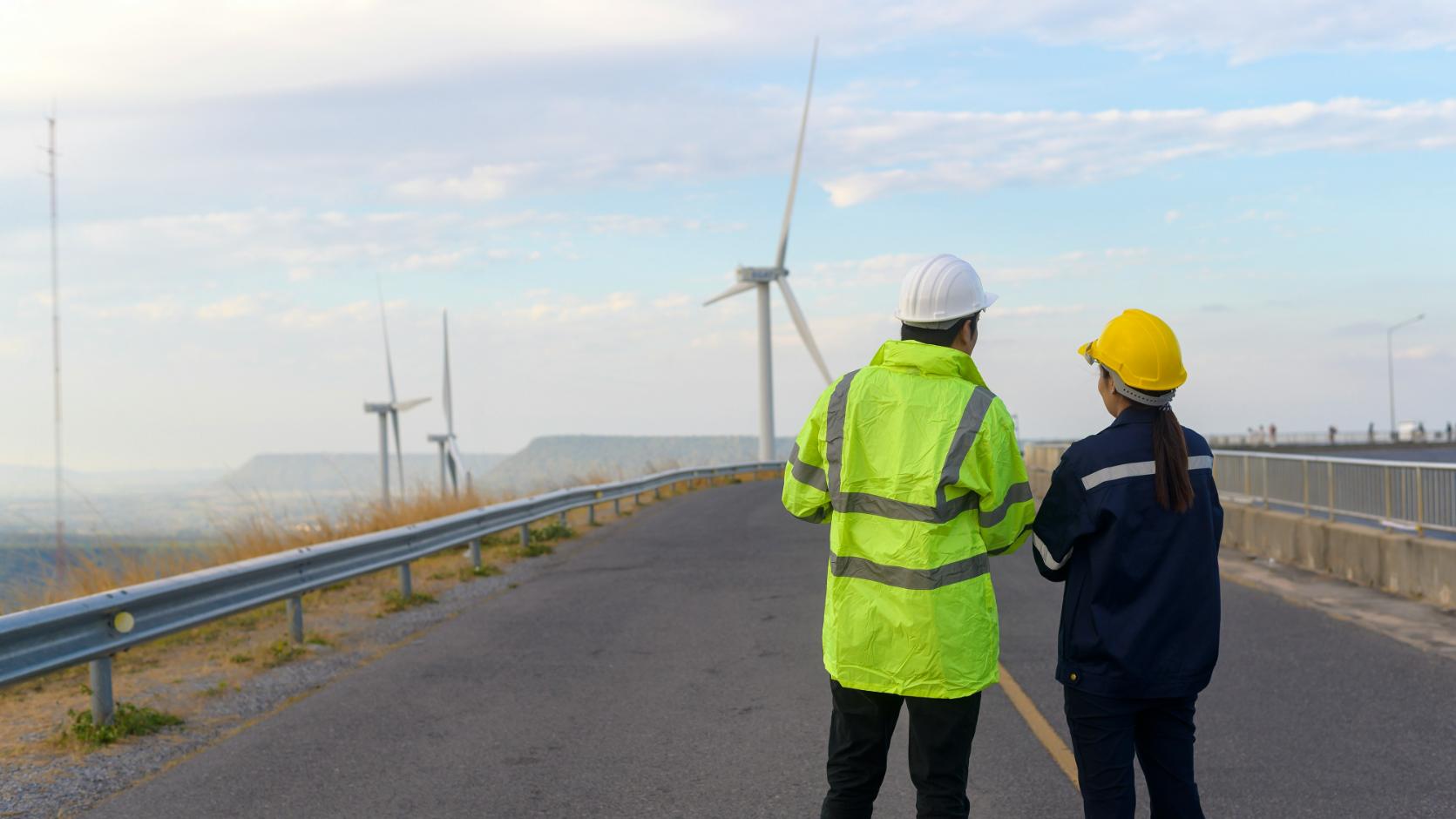 Zwei Personen in Warnwesten stehen auf Straße vor Windrädern