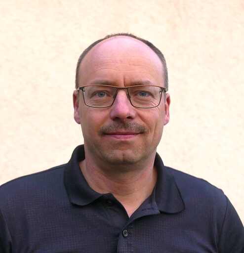 Porträt eines Mannes mittleren Alters mit Brille und dunklem Poloshirt vor hellem Hintergrund