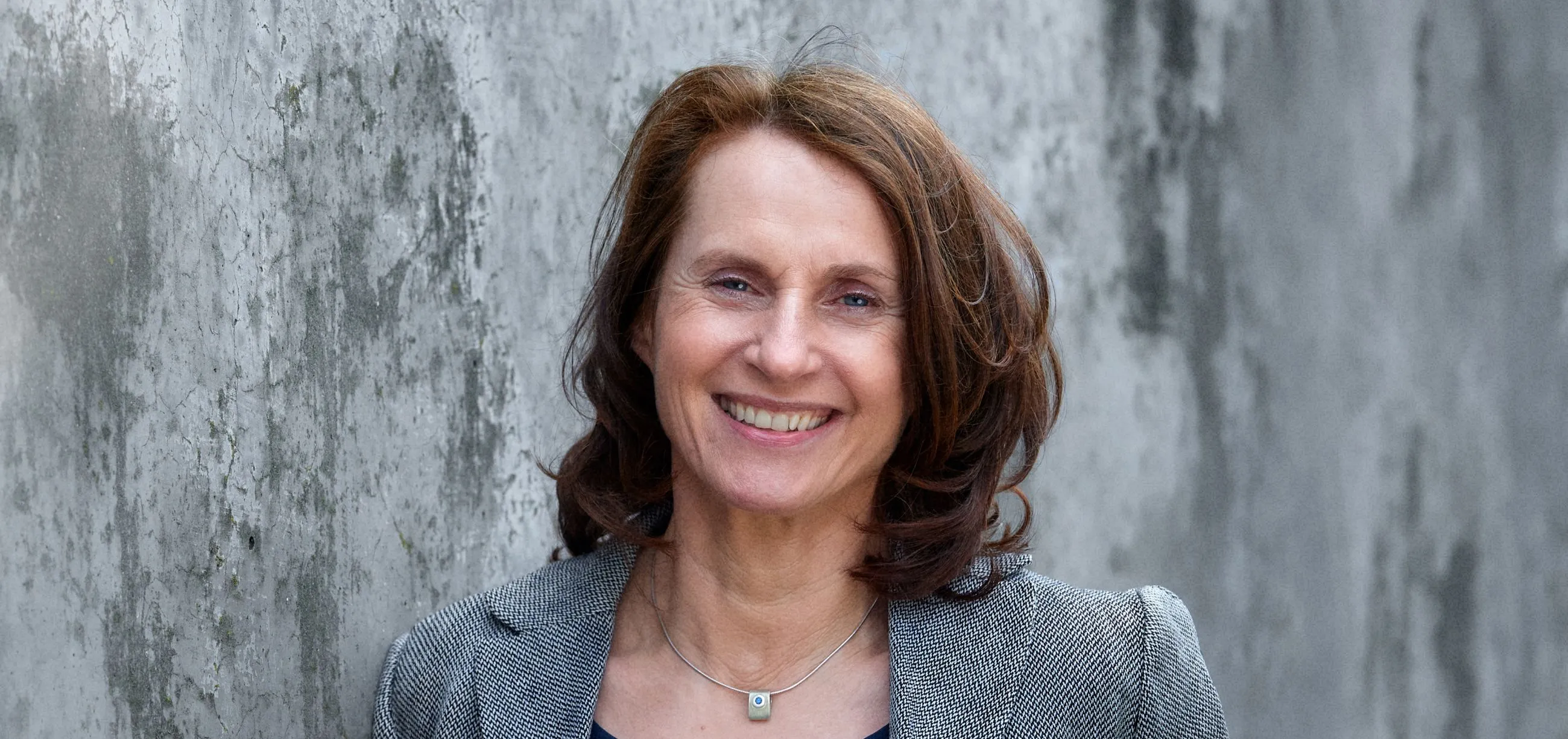 Frau in einem grauen Blazer vor einer Betonwand. 