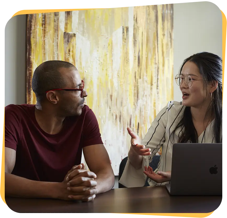 Two people sitting at a table engaged in conversation, with a laptop in front of the woman who is gesturing while talking.