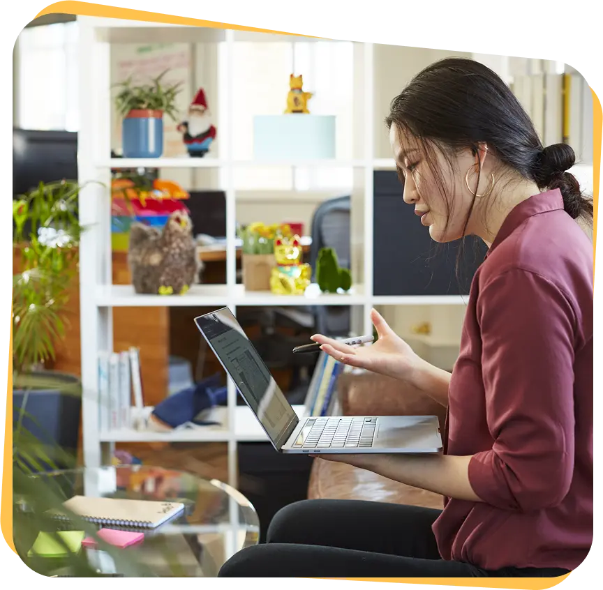 A woman in a burgundy shirt sits on a couch holding a laptop and gesturing with a pen during a video call in a cozy room with a white shelving unit behind her.