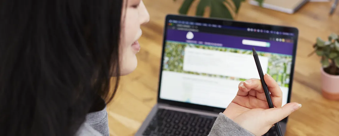Person holding a stylus pen in front of a laptop screen showing a webpage with purple header.