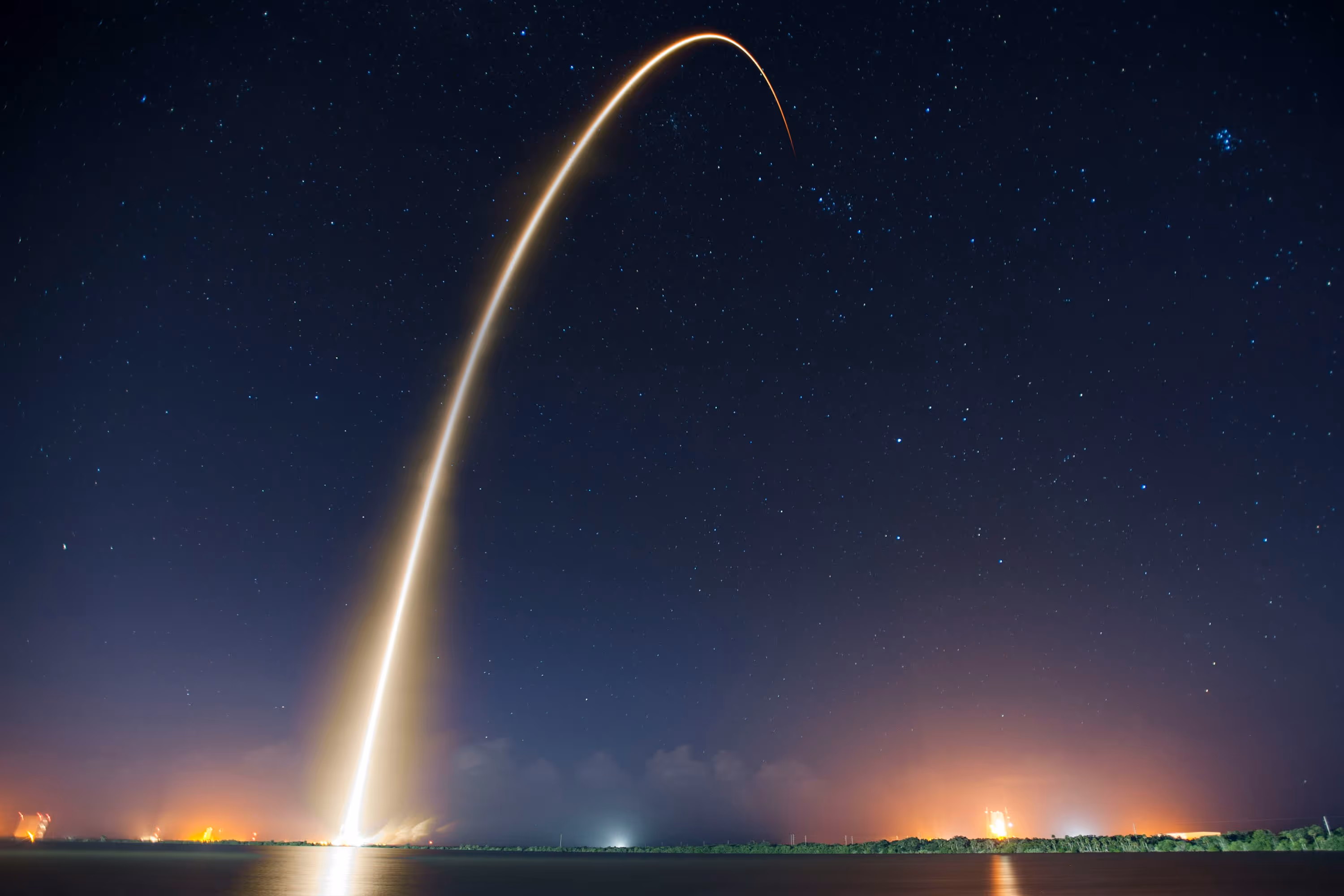 Long-exposure photo of a rocket launch at night creating a bright arc of light against a starry sky over water and distant land.
