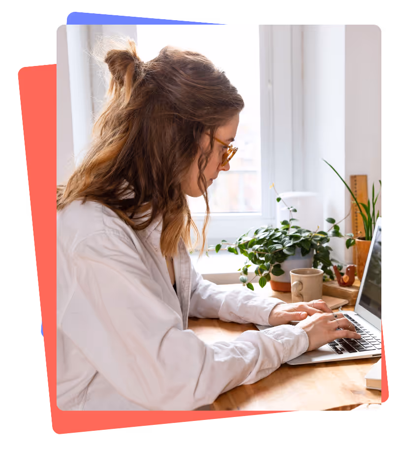 Woman with glasses typing on laptop at a wooden desk near a window with potted plants and a coffee mug.