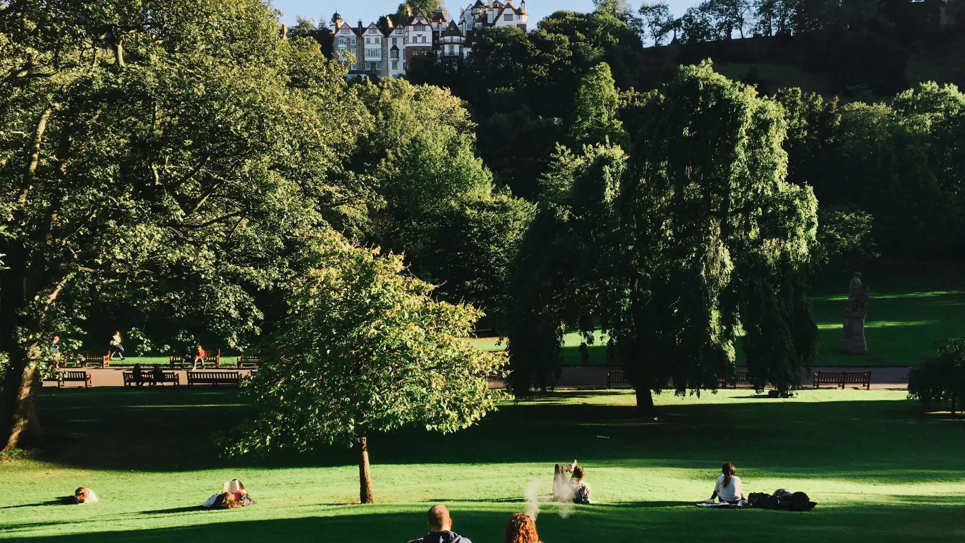 People sitting and relaxing on green grass in a sunny park with large trees and multi-story houses in the background.