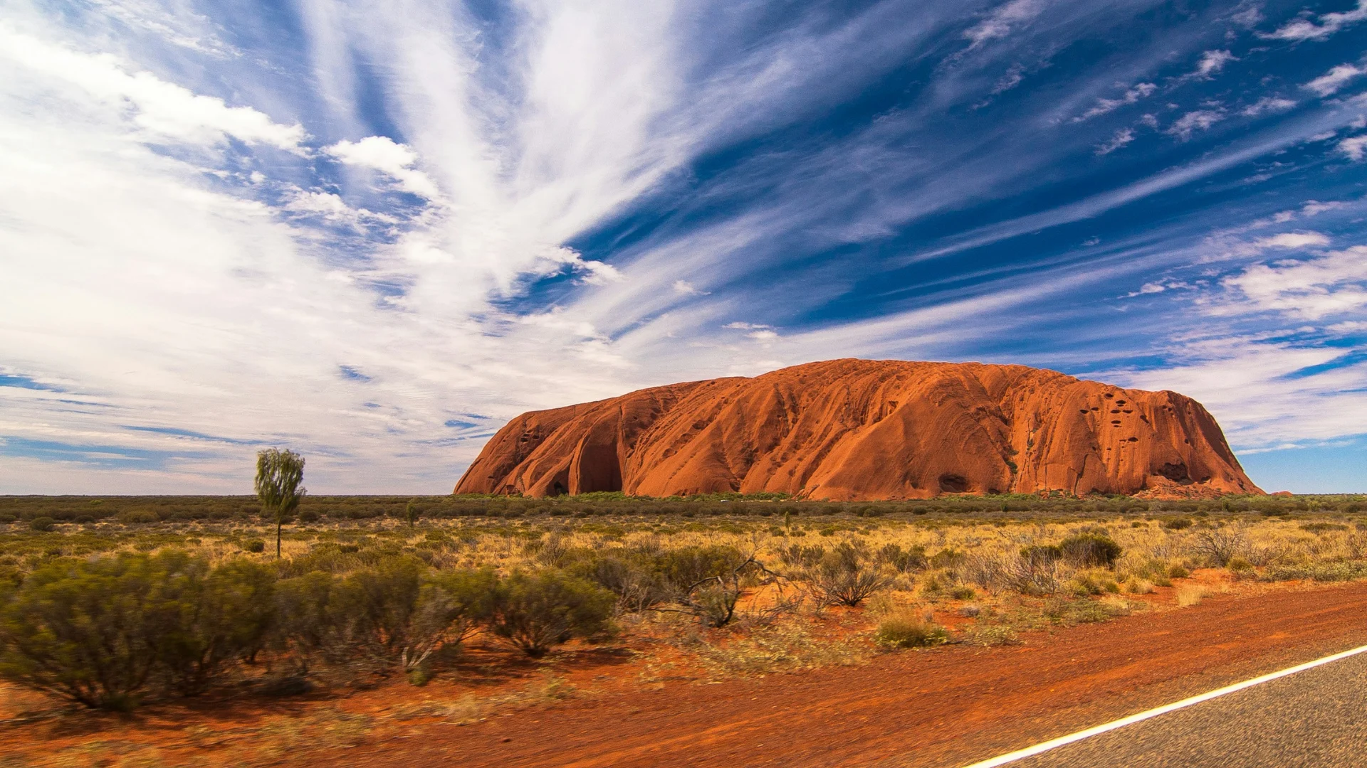 Uluru, a large red sandstone rock formation in a desert landscape under a blue sky with streaky white clouds.