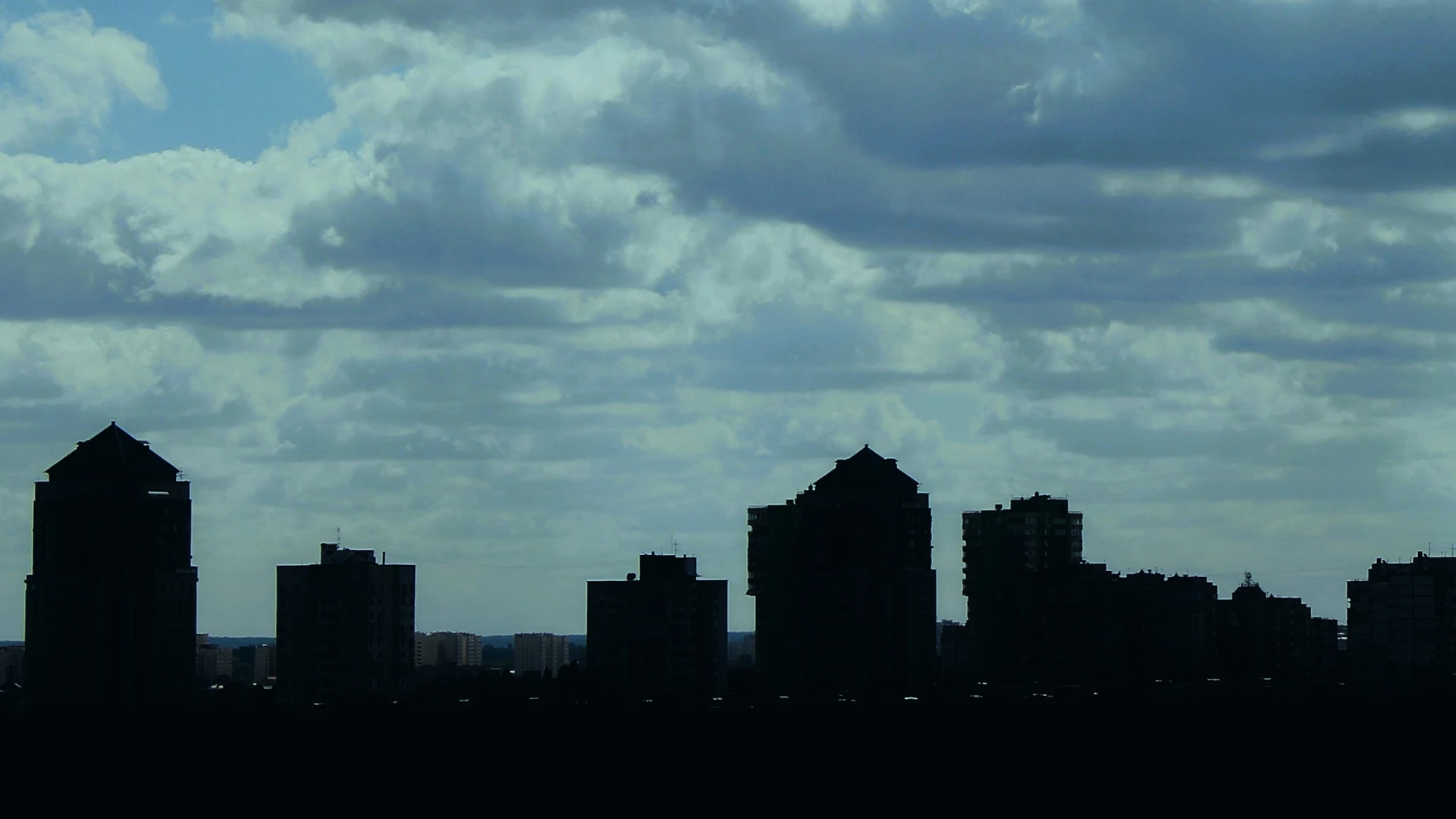 Silhouetted city skyline of various tall buildings under a cloudy sky.