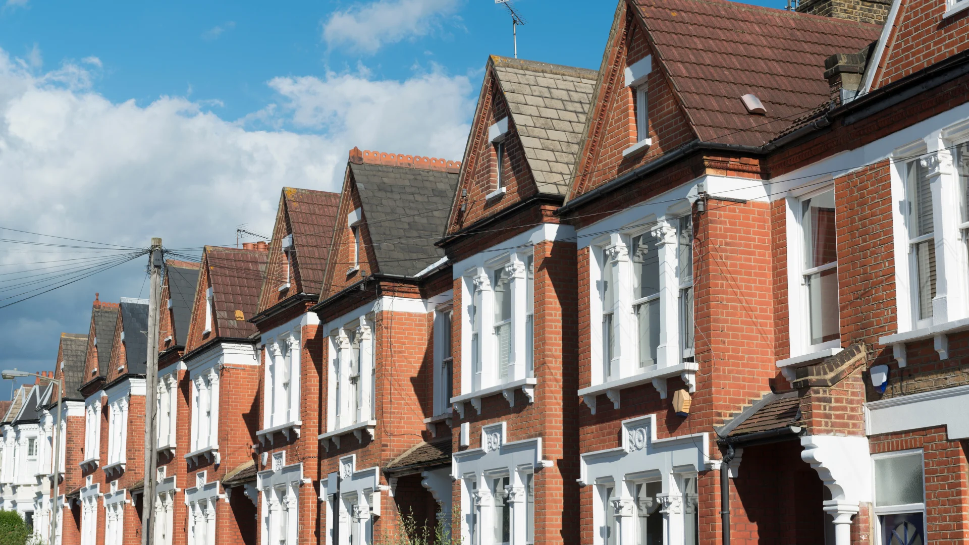Row of red brick terraced houses with white window frames and pitched roofs under blue sky with clouds.