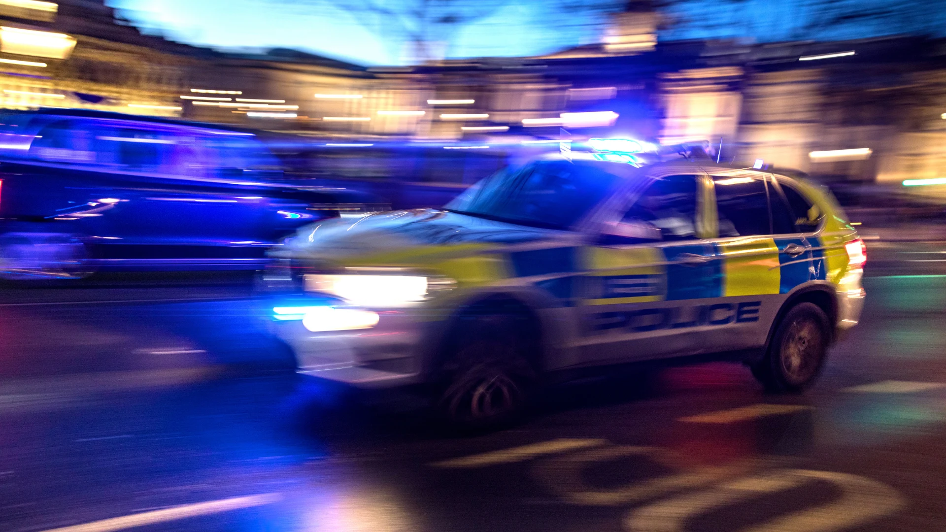 Blurred police car with flashing blue lights driving on a wet city street at dusk.