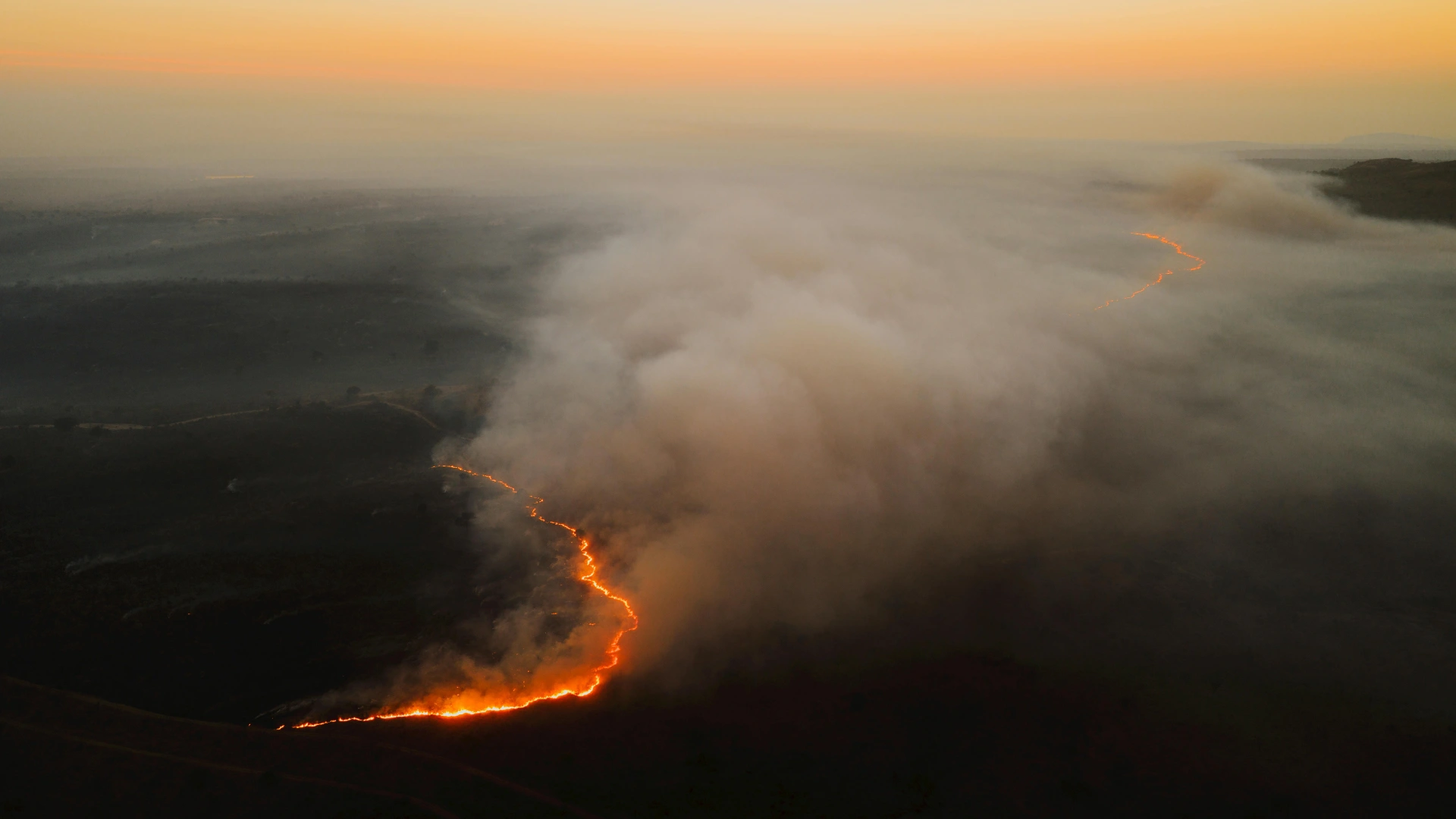 Aerial view of a wildfire burning through a dark landscape with thick smoke and an orange sky at sunset.
