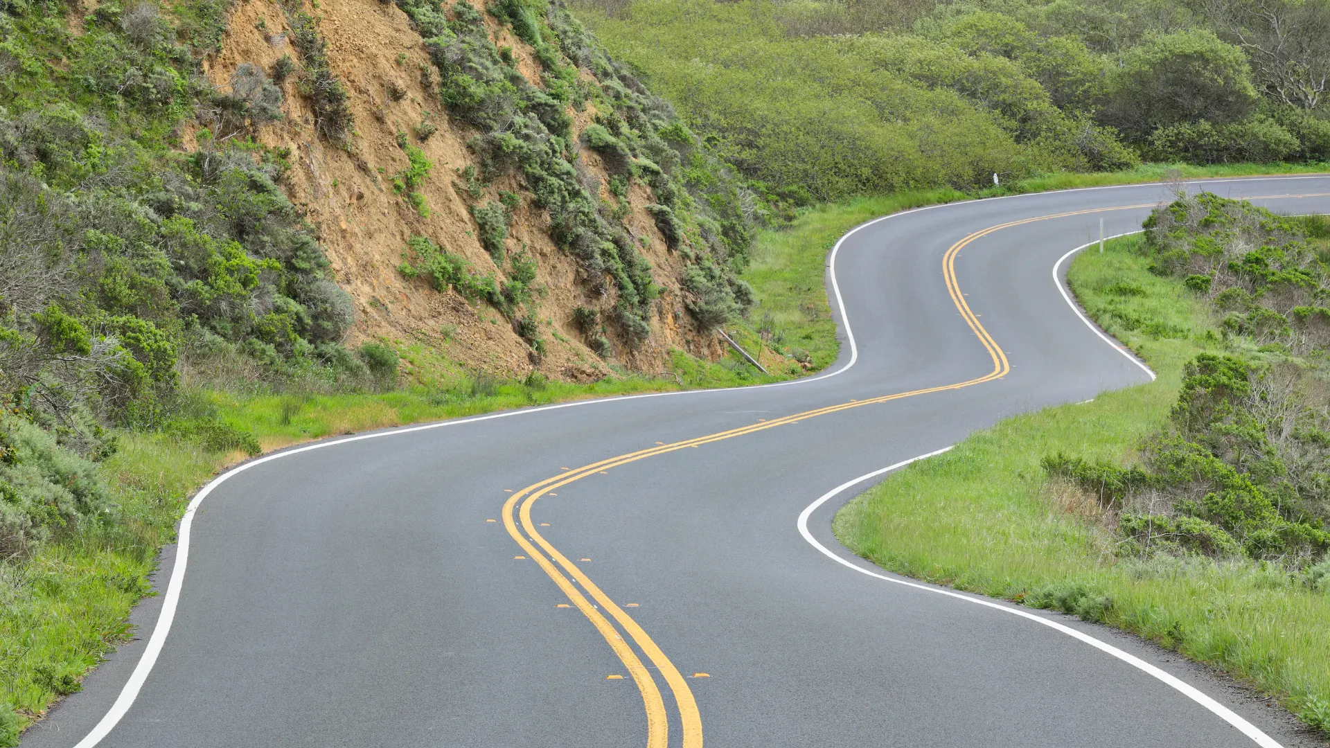 Curvy highway with double yellow lines running alongside green vegetation and a rocky hillside.